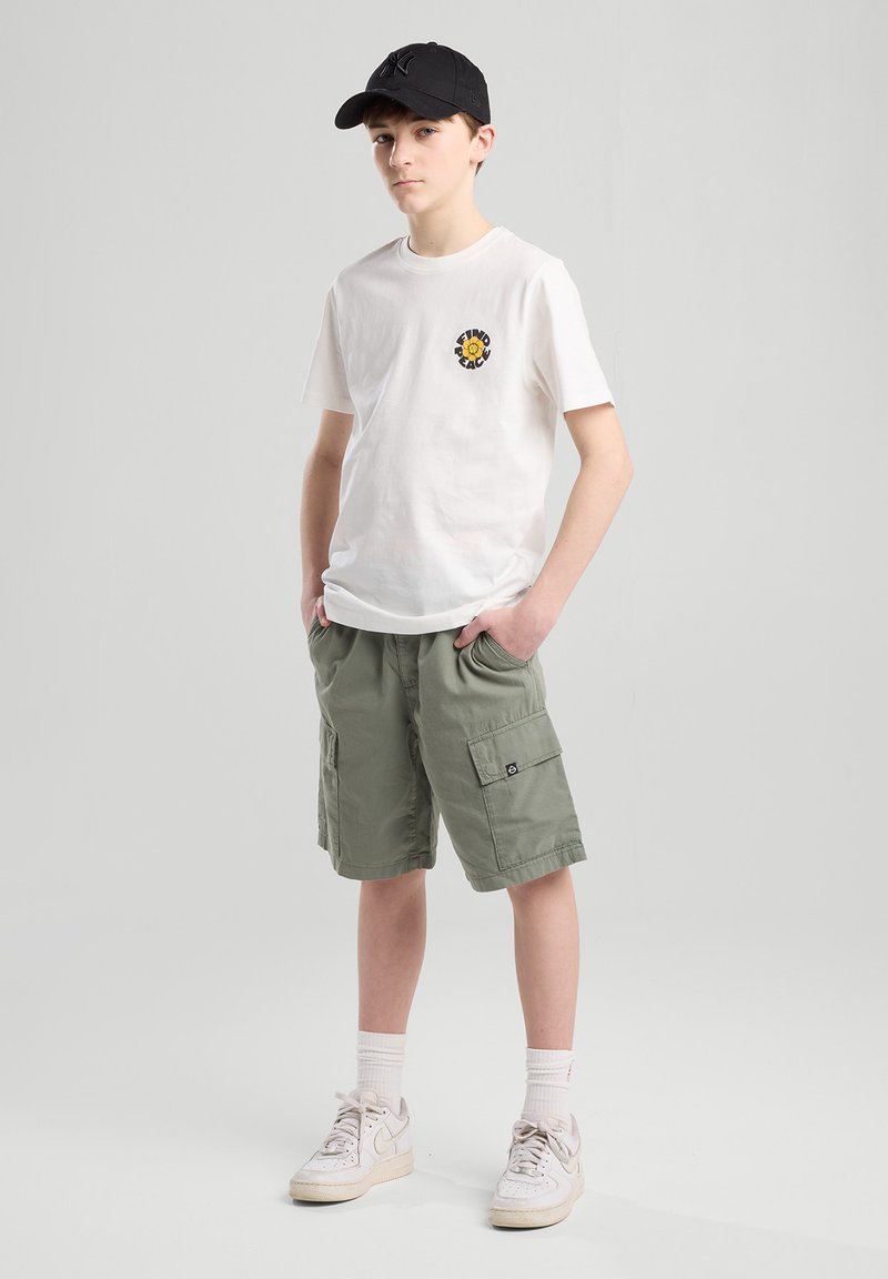T-shirt bianco in cotone con un logo circolare colorato, pantaloni cargo verde oliva, calze bianche e sneakers beige, abbinati a un cappellino nero.