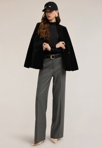 Schwarzer Samtblazer über einem transparenten schwarzen Oberteil, graue Hosen mit weitem Bein, schwarzer Gürtel mit goldener Schnalle und beigefarbene spitze High Heels. Schwarze Kappe mit Akzenten.