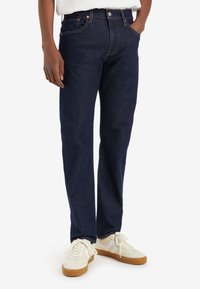 Mörkblå denimjeans med rak passform, fem fickor och kopparfärgade sömmar, kombinerat med vita sneakers med tan-färgade detaljer.