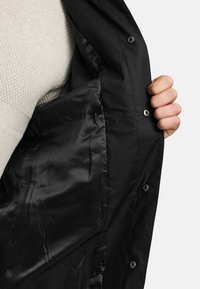 Musta ulkotakki, jossa on sileä satiinivuori. Siinä on painonappisulkimet ja sivutasku, ja sen ulkopinta on tekstuurillinen.