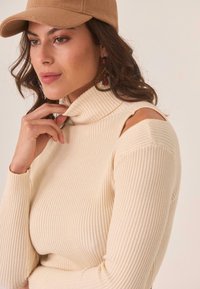 Suéter de punto de canalé color crema con diseño de hombros descubiertos, combinado con una gorra beige y grandes pendientes de gota con acentos rojos.