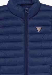 Granatowa kurtka puchowa z pionowym pikowaniem, wysokim kołnierzem i zapięciem na suwak z przodu. Na piersi znajduje się trójkątne logo marki.