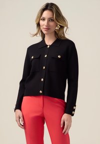 Cardigan nero con colletto, bottoni dorati e due tasche frontali. Abbinato a pantaloni rossi, con dettagli a bottoni sulle maniche.