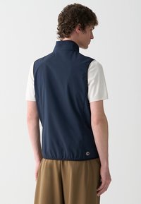 Gilet leggero blu navy con colletto alto, texture liscia e orlo inferiore elastico. Presenta un piccolo dettaglio del logo sul retro.