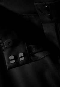 Dettaglio di indumento in tessuto nero che mostra un bottone rotondo e due clip metalliche rettangolari. Presenta una texture liscia e cuciture nascoste.