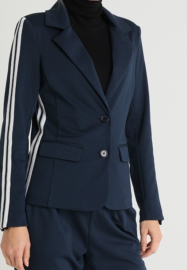Blazer bleu marine avec deux boutons à l'avant et des rayures blanches sur les manches, porté sur un col roulé noir et un pantalon bleu marine assorti.