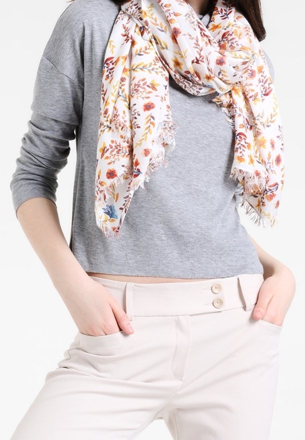 Femme portant un haut gris à manches longues, un pantalon crème avec double boutonnage, et une écharpe blanche fleurie avec des bords frangés. Mains dans les poches.