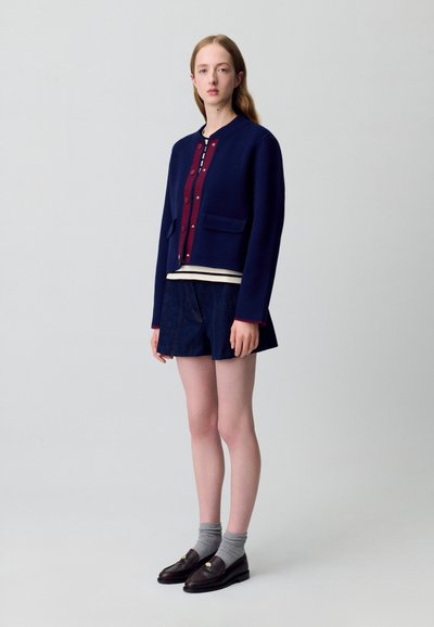 Giacca cropped blu navy con bottoni bordeaux, due tasche anteriori. Indossata sopra una maglietta a righe e pantaloni corti in denim. Mocassini scuri con calzini grigi.