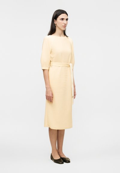 Γυναίκα με σκούρα μαλλιά που φοράει αμυγδαλί φόρεμα μήκους μέχρι το γόνατο με μανίκια 3/4, ζώνη στη μέση και σκούρα επίπεδα παπούτσια, στέκεται μπροστά σε ένα μονόχρωμο φόντο.