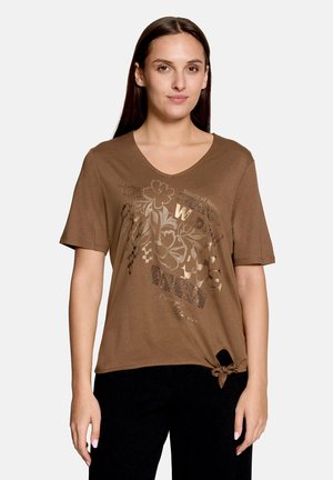 Femme portant un t-shirt marron à col en V avec des motifs floraux et des textes, et un détail de nœud noué à l'ourlet, associé à un pantalon noir.