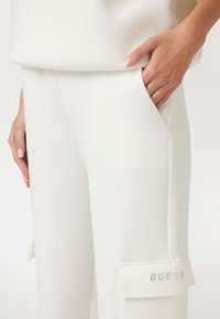 Bílé joggingové kalhoty vyrobené z měkké tkaniny, s boční kapsou a nášivkou s nápisem "GUESS" v perforovaném designu.
