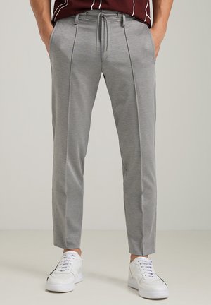 Homme portant un pantalon gris ajusté avec cordon de serrage et des baskets blanches, les mains dans les poches, debout sur un sol clair devant un fond uni.