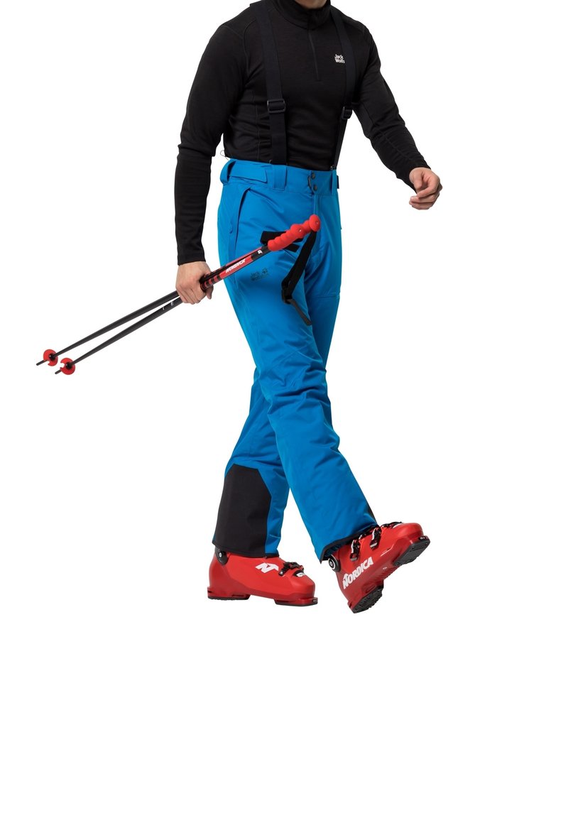 Homme portant un pantalon de ski bleu, des chaussures de ski rouges, un haut noir et des bretelles, tenant des bâtons de ski rouges et noirs en marchant.