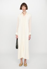 Largo vestido de color crema con un cuello, frente con pliegues, mangas largas y puños en contraste. La modelo sostiene un bolso negro texturizado y lleva zapatos planos dorados.