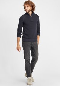 Tumesinine pullover pool tõmblukuga kraega, tekstuuriga kudumise muster, soonikkoes mansetid ja äär, koos tumehallide teksade ja heledate kingadega.