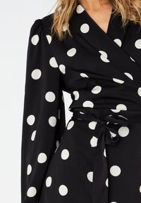 Vestito nero avvolgente con pois bianchi, maniche lunghe e vita legata. Il tessuto ha una texture liscia con una finitura opaca.