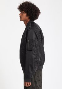 Giubbotto bomber nero con polsini a costine, tasca con zip sulla manica e superficie liscia. Presenta una vestibilità rilassata e un design minimalista.