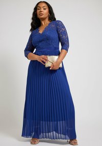 En lång, plisserad aftonklänning i rik blå färg. Spetstopp med trekvartsärmar och en djup V-ringning. Klasad med en liten gyllene kuvertväska.