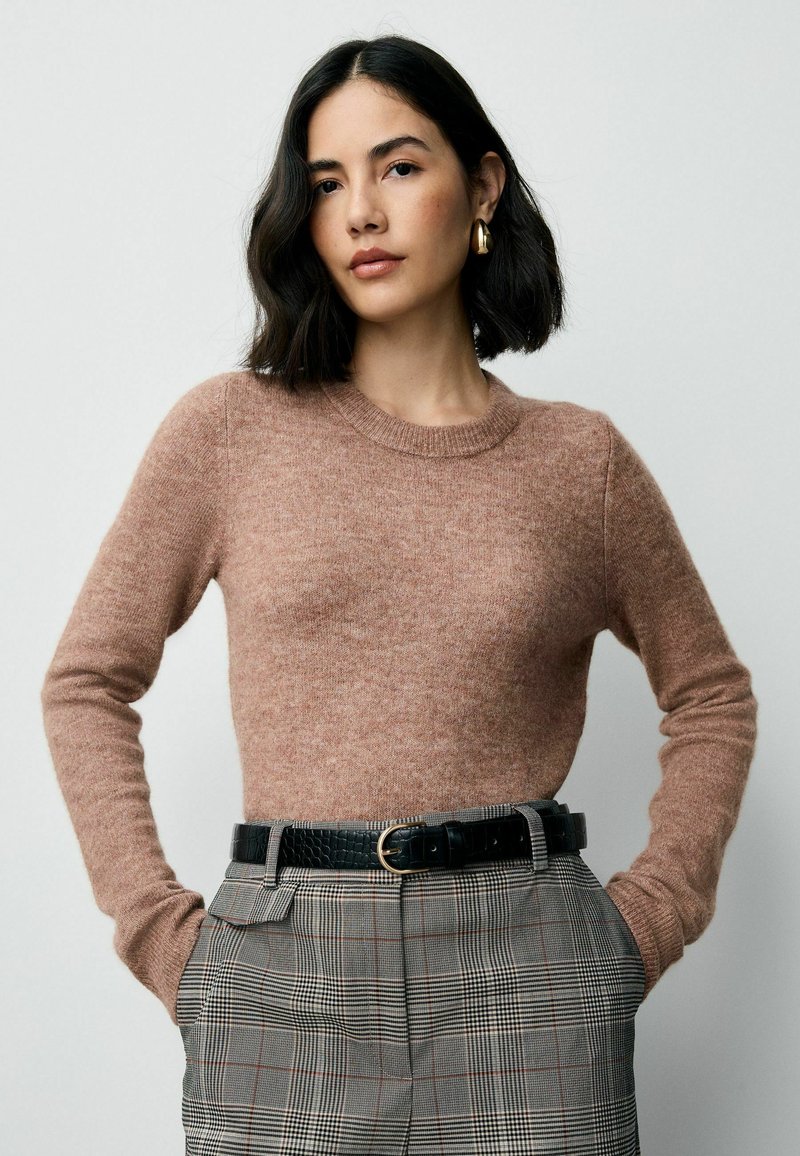 Hellbrauner, gestrickter Pullover mit langen Ärmeln und Rundhalsausschnitt, kombiniert mit karierter Hose und einem schwarzen Gürtel. Angenehme Textur.