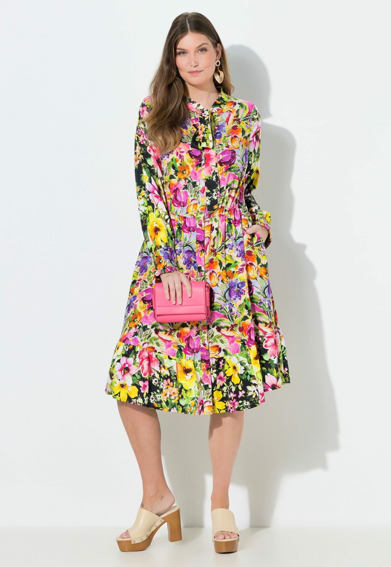 Frau trägt ein buntes, florales Midikleid, beige Plateau-Pumps, hält eine kleine pinke Handtasche und trägt herzförmige Ohrringe.