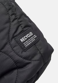 Tissu matelassé noir avec une texture brillante ; comporte une étiquette rectangulaire indiquant "RECYCLÉ" et des détails sur le produit.