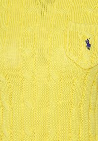 Gul stickad tröja med ett texturerat snöre-mönster. Har en liten ficka och en lila logotyp. Materialet verkar mjukt och av medelvikt.