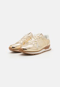 Gullsneakers i metallisk gull med en glatt overflate, snøring, bred tunge og brun gummisåle. Detaljer inkluderer en hvit kant og logo.