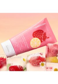 Un tubetto rosa di Ole Henriksen Strawberry Lemonade Scrub con gocce di spray, caratterizzato da grafiche di fragola e limone, angolato sopra cubetti di ghiaccio con fragole.