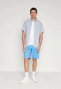 Mann står med en lyseblå kortermet skjorte over en hvit t-skjorte, blå shorts, hvite sokker og hvite joggesko.