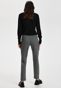 Femme debout de dos portant un haut noir à manches longues, un pantalon gris tailleur avec poches arrière, et des chaussures noires épaisses sur fond blanc.