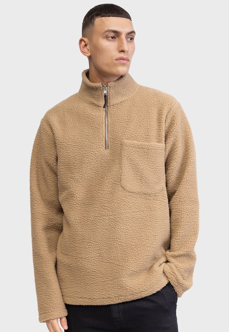 Beige fleecegenser med høy krage, halv-lukkning og brystlomme. Den har teksturert stoff og lange ermer.