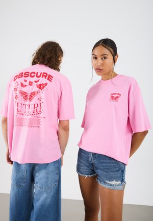 Pink cotton t-shirts feature a bold red graphic design with text and butterfly motifs. One shirt has short sleeves; the other is oversized.