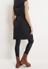 Gilet imbottito trapuntato nero con cappuccio, fascia elastica in vita, indossato su una maglietta a maniche lunghe beige a coste, abbinato a leggings neri simili alla pelle e stivali alla caviglia marroni.