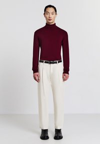 Bordeauxrotes Rollkragenoberteil mit langen Ärmeln, kombiniert mit cremefarbenen plissierten Hosen, ergänzt durch einen schwarzen Gürtel und Stiefeletten.