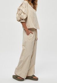 Beige gestreifter Crop-Top mit puffärmel, kombiniert mit weit geschnittenen Hosen. Strukturierter Stoff, seitliche Taschen und olivgrüne Sandalen.