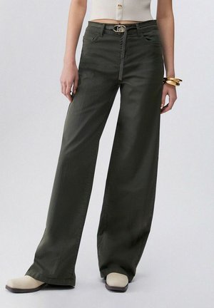 Donna che indossa pantaloni verde scuro a vita alta e gamba larga con cintura, top bianco corto, scarpe beige con punta quadrata e braccialetti d'oro a un polso.