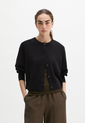 Vrouw met lang bruin haar gebonden draagt een zwart cardigan met knopen over een olijfgroene top en losse olijfgroene broek, staand met handen in de zakken.