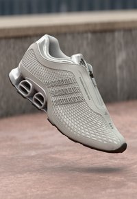 Zapatilla deportiva de color gris claro con parte superior texturizada, cierre de cremallera y suela acolchada que presenta tres cortes semi-circulares para soporte.
