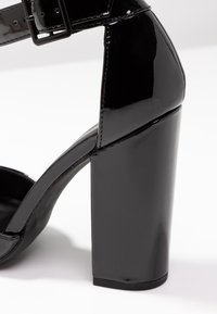 Sandale à talons hauts en cuir verni noir avec un large talon bloc, une bride de cheville et une finition lisse et brillante.