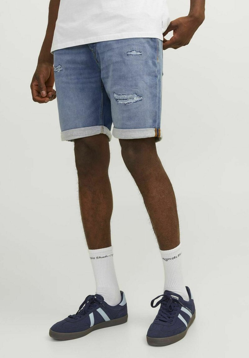 Pantalones cortos de mezclilla azul claro con parches deshilachados, puños enrollados y un ajuste ligeramente holgado. Llevado con zapatillas de deporte azul marino y calcetines blancos.