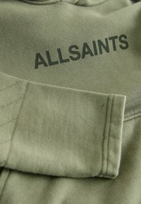 Tkanina bluzy w oliwkowej zieleni o teksturowanej powierzchni. Z napisem „ALLSAINTS” w ciemnozielonym kolorze oraz widocznym przeszyciem wzdłuż rękawów.