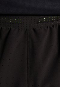 Svarta atletiska shorts med en perforerad midja som har små gröna accenter, tillverkade av ett slätt, lätt tyg, designade för ventilation.