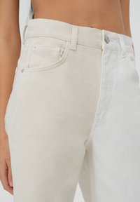 Jeans de mezclilla de color claro con un diseño bicolor que presenta beige y blanco. Los detalles incluyen un cierre de botón y cremallera, bolsillos laterales y costuras visibles.
