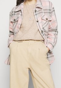 Rosa- und graue karierte Jacke mit metallischen Knöpfen, über einem beigen strukturierten Pullover und beigen Hosen mit Faltendetails geschichtet.