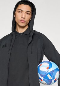 Μαύρο φούτερ με φερμουάρ και κουκούλα με κορδόνια, φορεμένο πάνω από μαύρο πόλο πουκάμισο. Άτομο κρατά μια πολύχρωμη μπάλα ποδοσφαίρου με κυματιστά σχέδια.