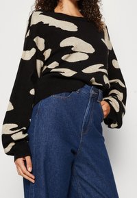 Schwarzer Pullover mit beigem abstrakten Muster, lockere Passform, gerippte Bündchen und Saum. Kombiniert mit hochgeschnittenen blauen Denim-Hosen.