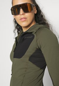 Olive green zip-up jacket with black panels, textured fabric, adjustable hood, and high neckline. Large orange-tinted sunglasses, rectangular shape.