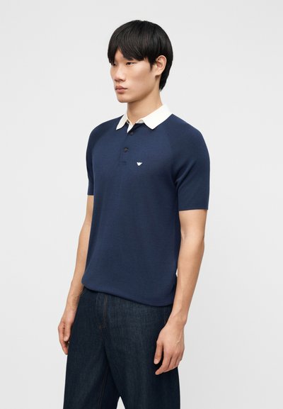 Νεαρός άνδρας που φοράει μπλε κοντομάνικο πόλο με λευκό γιακά και σκούρο τζιν, στέκεται μπροστά σε απλό φωτεινό φόντο.