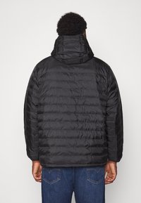 Man som bär en svart vadderad huva jacka och blå jeans, står vänd bortåt mot en enfärgad ljus bakgrund.