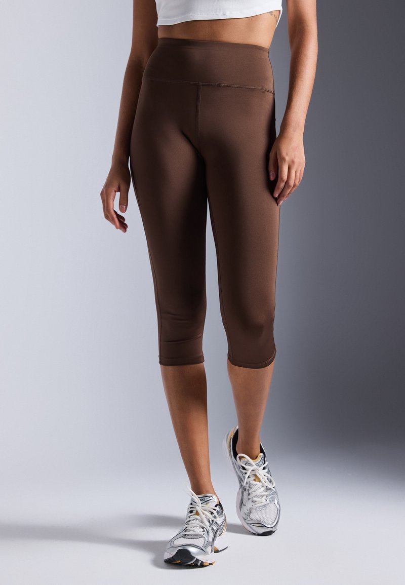 Femme portant des leggings capri marron taille haute et des chaussures de course blanches, debout sur un fond gris uni.